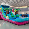 Large tropical-themed inflatable water slide featuring a single blue sliding lane, palm tree decorations, colorful flower accents, and an extended splash pool with vibrant blue and pink base in indoor warehouse setting.