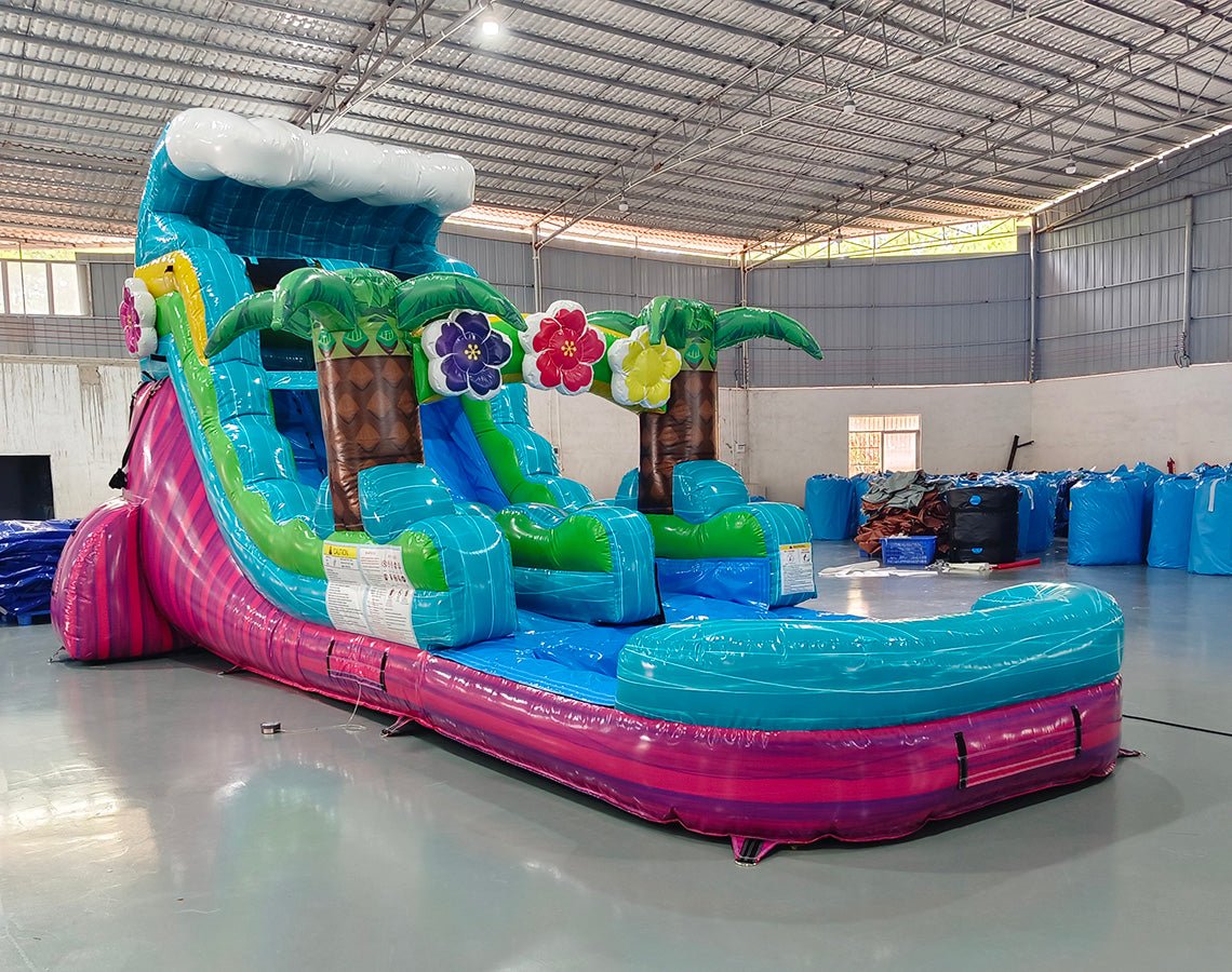 Large tropical-themed inflatable water slide featuring a single blue sliding lane, palm tree decorations, colorful flower accents, and an extended splash pool with vibrant blue and pink base in indoor warehouse setting.