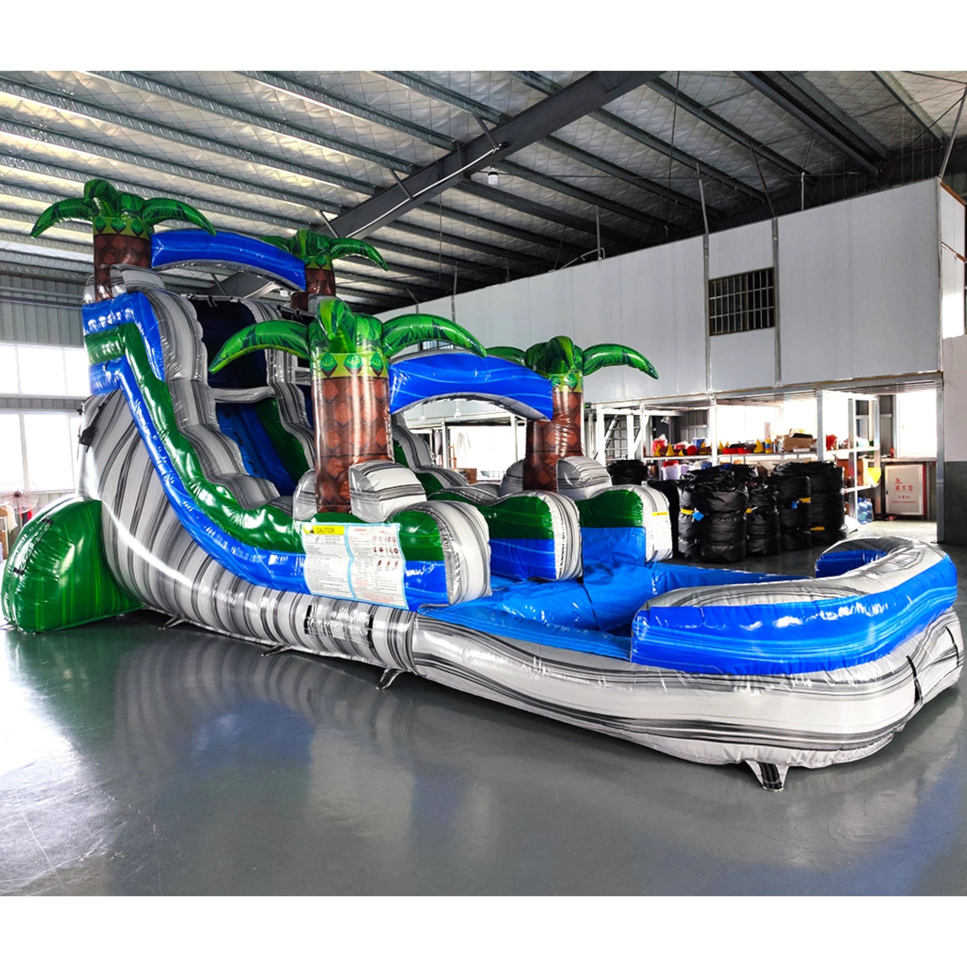 Large tropical-themed inflatable water slide featuring a single blue sliding lane, palm tree decorations, and an extended splash pool with blue and white color scheme.