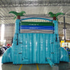 Rear view of large inflatable Bahama Blast water slide with turquoise base, three blue sliding lanes, palm tree decorations on top, and a central climbing structure, set up in an indoor warehouse.