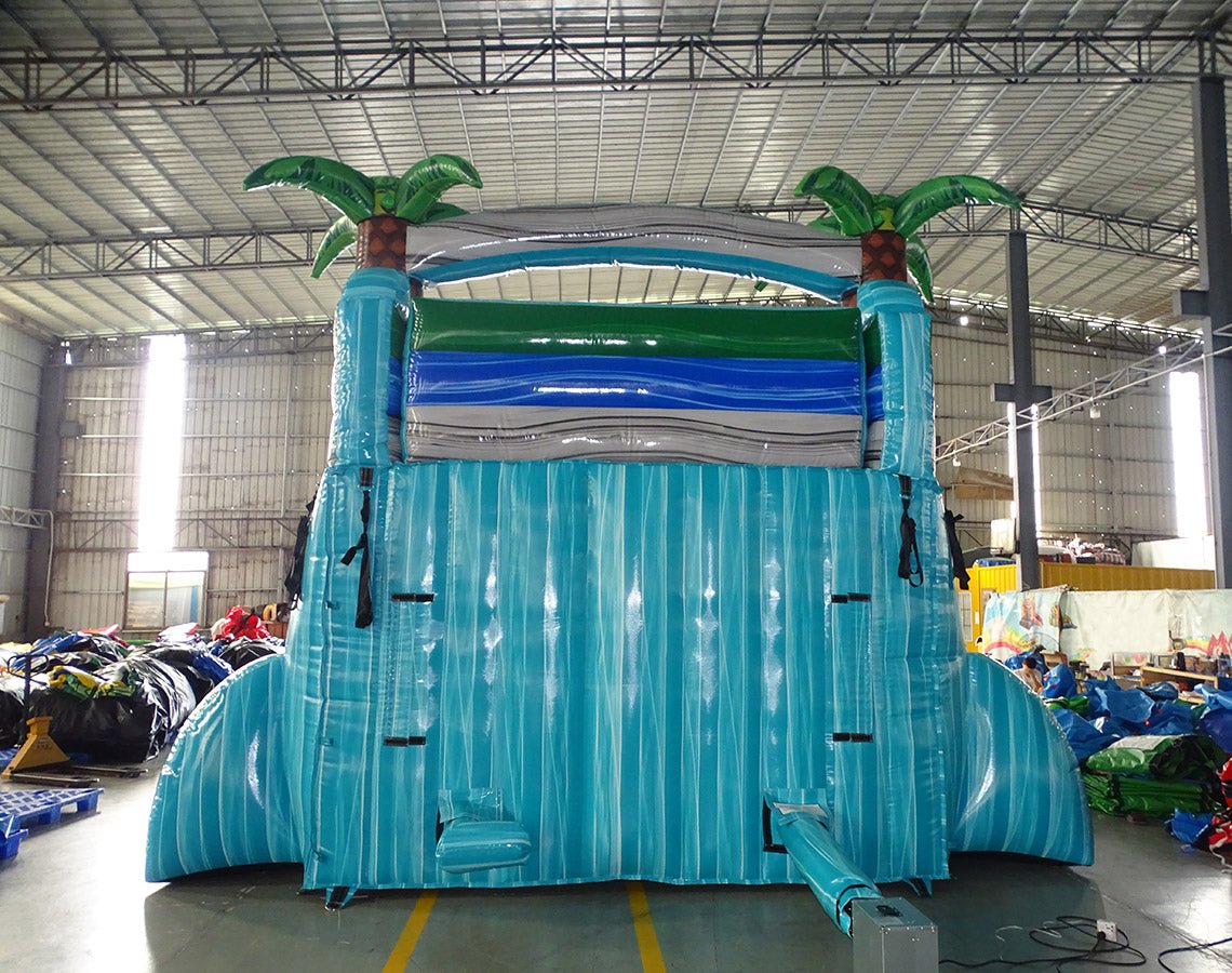 Rear view of large inflatable Bahama Blast water slide with turquoise base, three blue sliding lanes, palm tree decorations on top, and a central climbing structure, set up in an indoor warehouse.