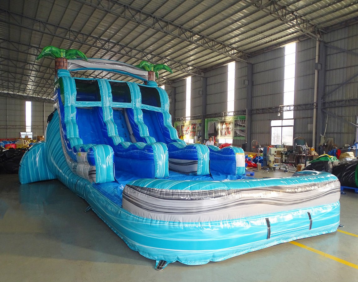 Close-up view of the 15ft Bahama Blast water slide displaying details of the blue sliding lanes, central climbing section, inflatable palm trees, and wave-patterned splash pool in an indoor facility.