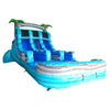 Large inflatable tropical-themed water slide with three blue lanes, palm tree decorations, and a center climbing wall, set up in an indoor warehouse space with colorful inflatables in the background.