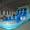 Side angle view of the Bahama Blast inflatable water slide featuring three blue lanes, central climbing structure, palm tree decorations, and extended splash area in a warehouse setting.