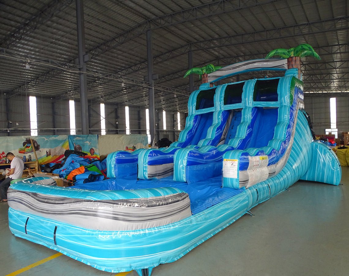 Side angle view of the Bahama Blast inflatable water slide featuring three blue lanes, central climbing structure, palm tree decorations, and extended splash area in a warehouse setting.