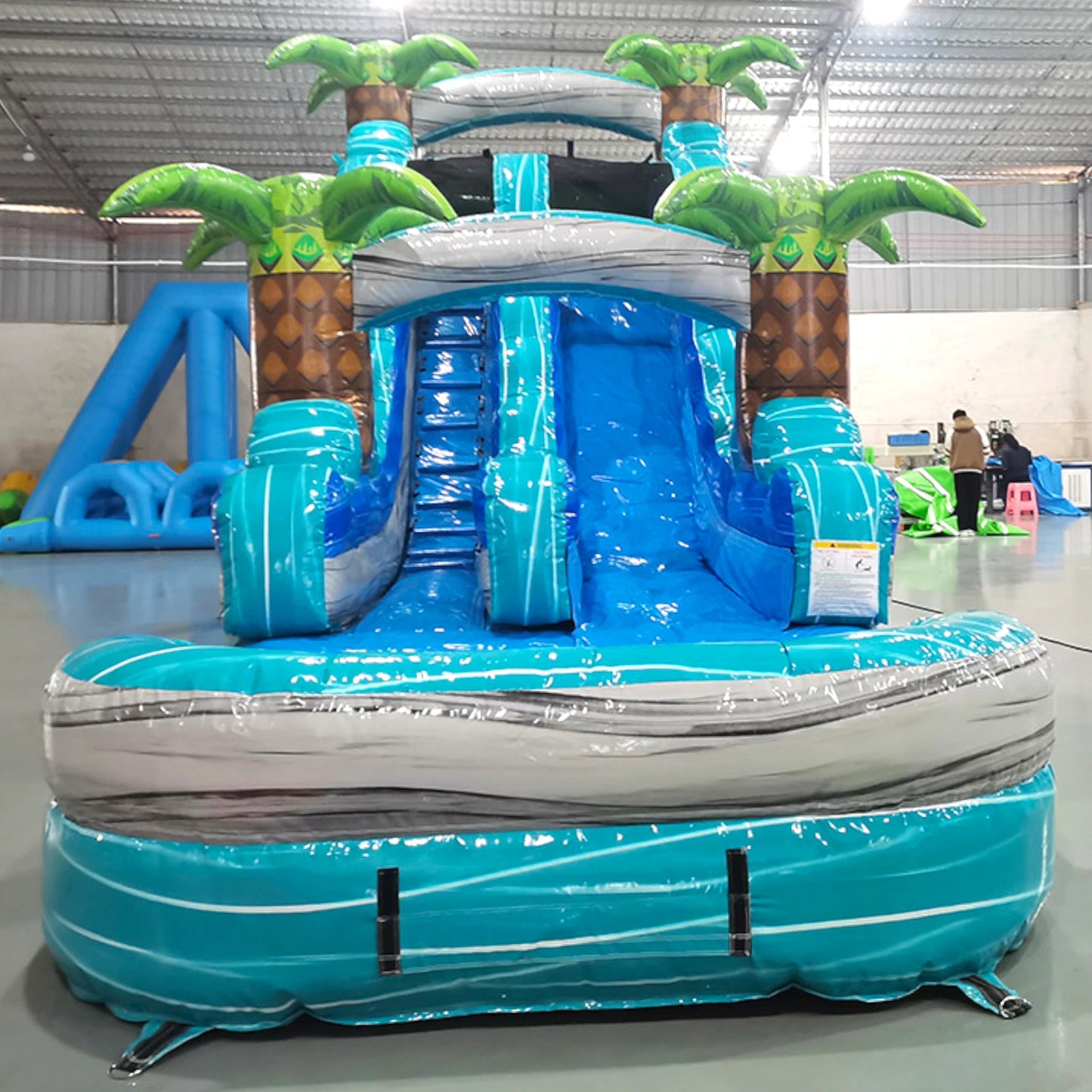 Front view of Bahama Blast single lane water slide in indoor setting, showing tall structure with blue sliding surface, palm trees, and large splash area.