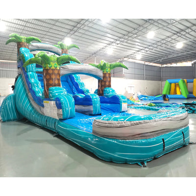 Large tropical-themed inflatable water slide featuring a single blue sliding lane, palm tree decorations, and an extended splash pool with turquoise and blue color scheme.