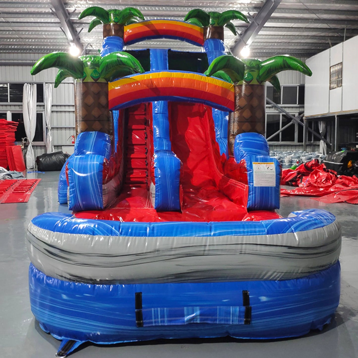 Front view of Baja Splash single lane water slide in indoor facility, showing vibrant blue and red design, palm tree decorations, and extended splash pool area with inflatable barriers.