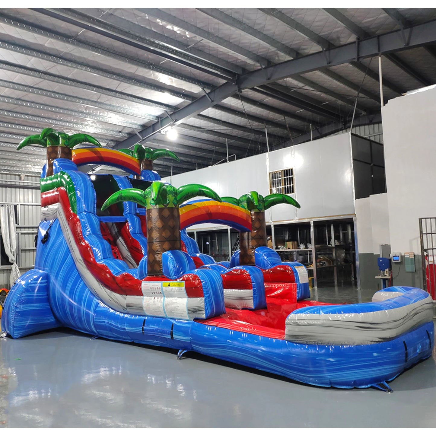 Large tropical-themed inflatable water slide featuring a single blue sliding lane, palm tree decorations, and an extended splash pool with vibrant blue and red colors in indoor warehouse setting.