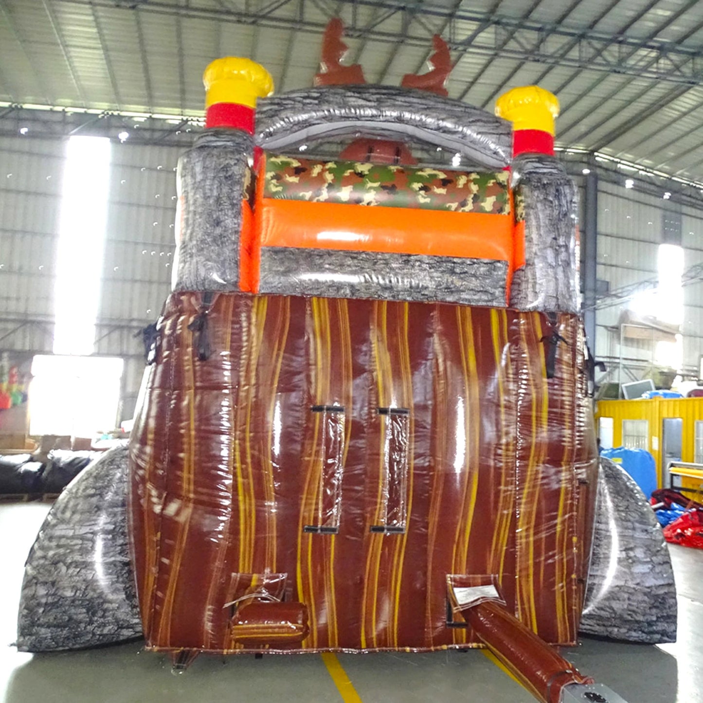 Rear view of Buck Shot single lane water slide in indoor warehouse, showing tall structure with tree bark texture, camouflage accents, and yellow safety caps on top.