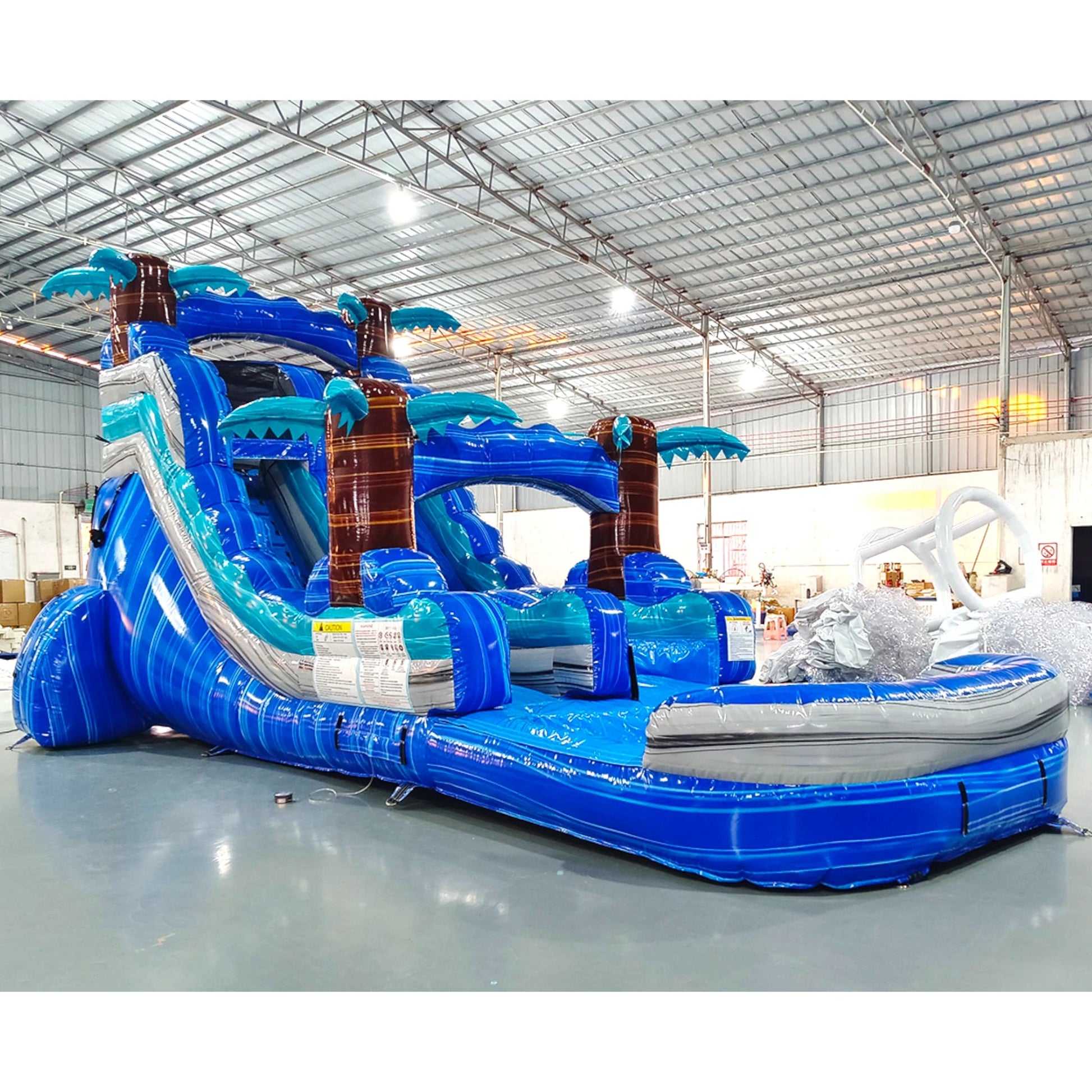 Side view of Cayman Crush single lane water slide showing full length with blue sliding surface, palm tree accents, and large splash area with tropical wave patterns.