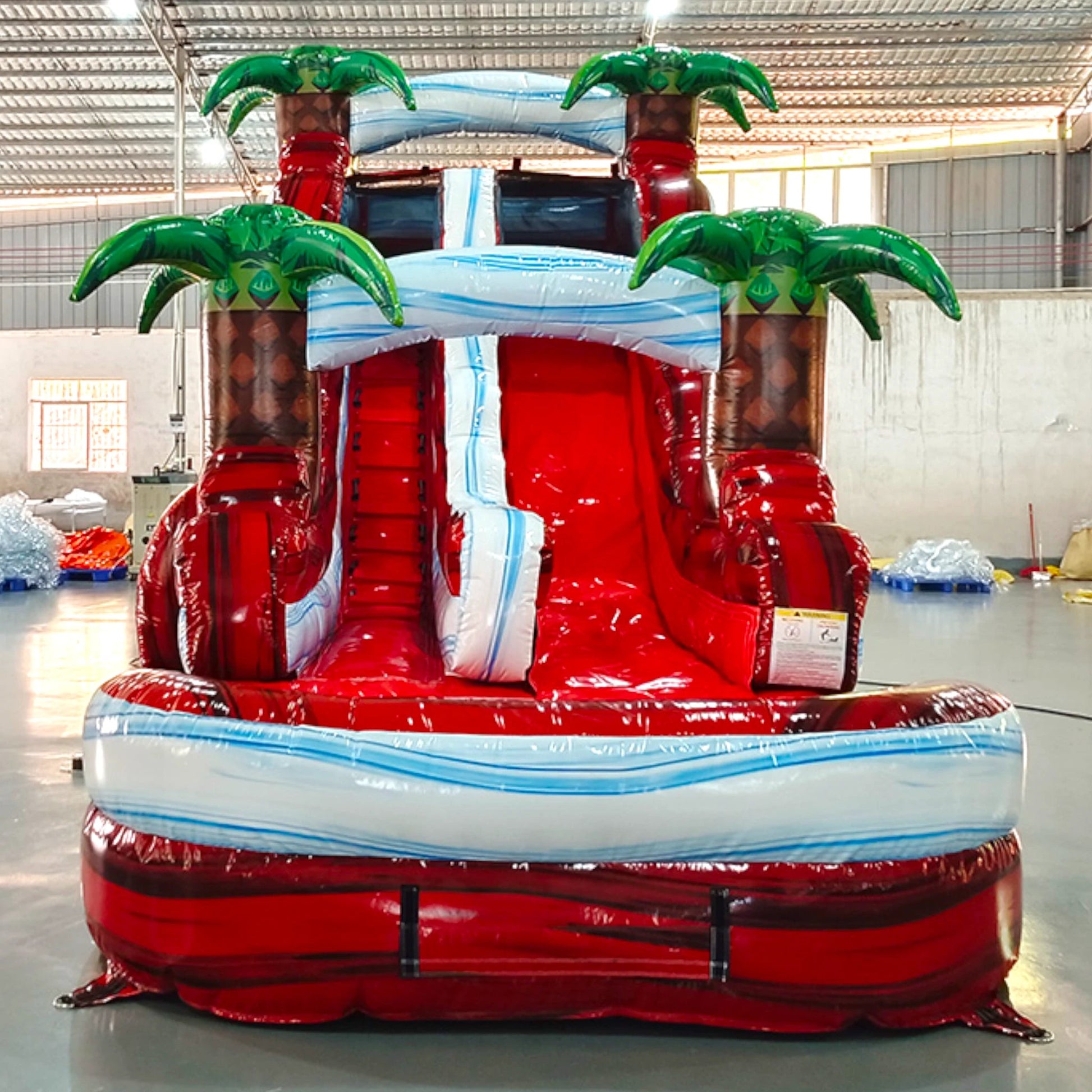 Front view of Crimson Bay single lane water slide in indoor warehouse, showing tall structure with red sliding surface, palm trees, and large splash area with blue accents.