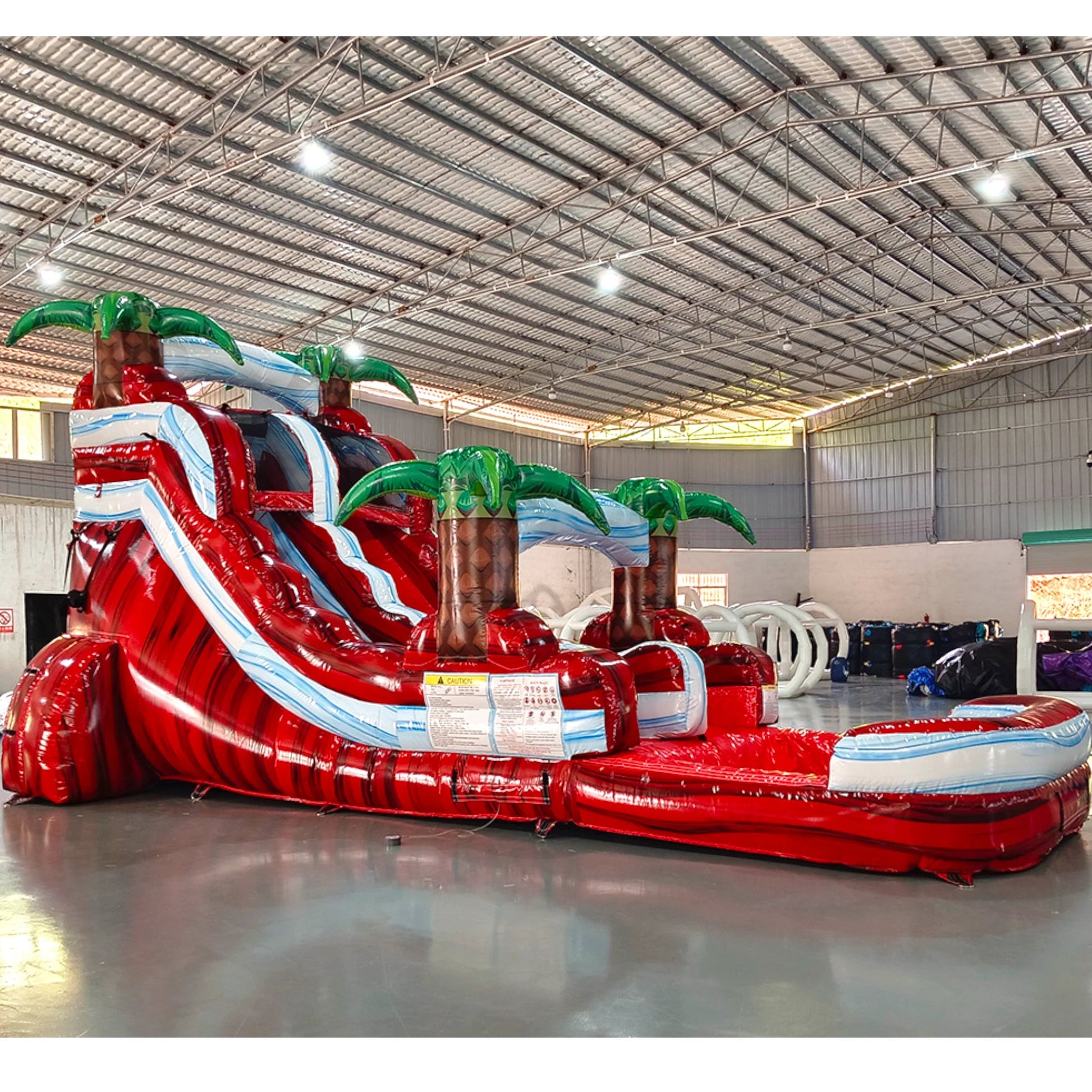 Large tropical-themed inflatable water slide featuring a single red sliding lane, palm tree decorations, and an extended splash pool with blue wave-like design.