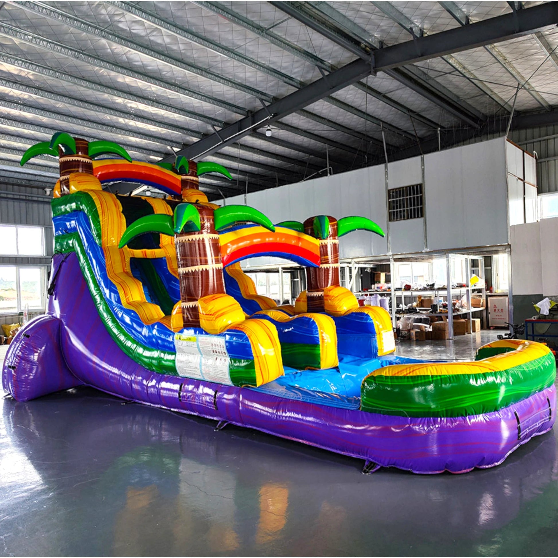 Side view of Goombay Splash inflatable water slide showing full length with blue sliding lane, palm trees, and extended splash pool in warehouse environment.