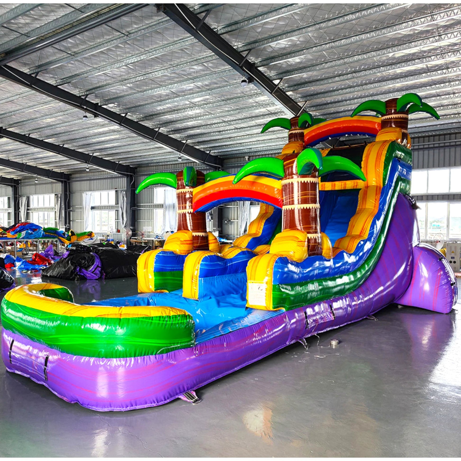 Angled view of Goombay Splash single lane water slide in indoor warehouse, showcasing its height, blue sliding surface, palm tree decorations, and extended purple and green splash pool area.