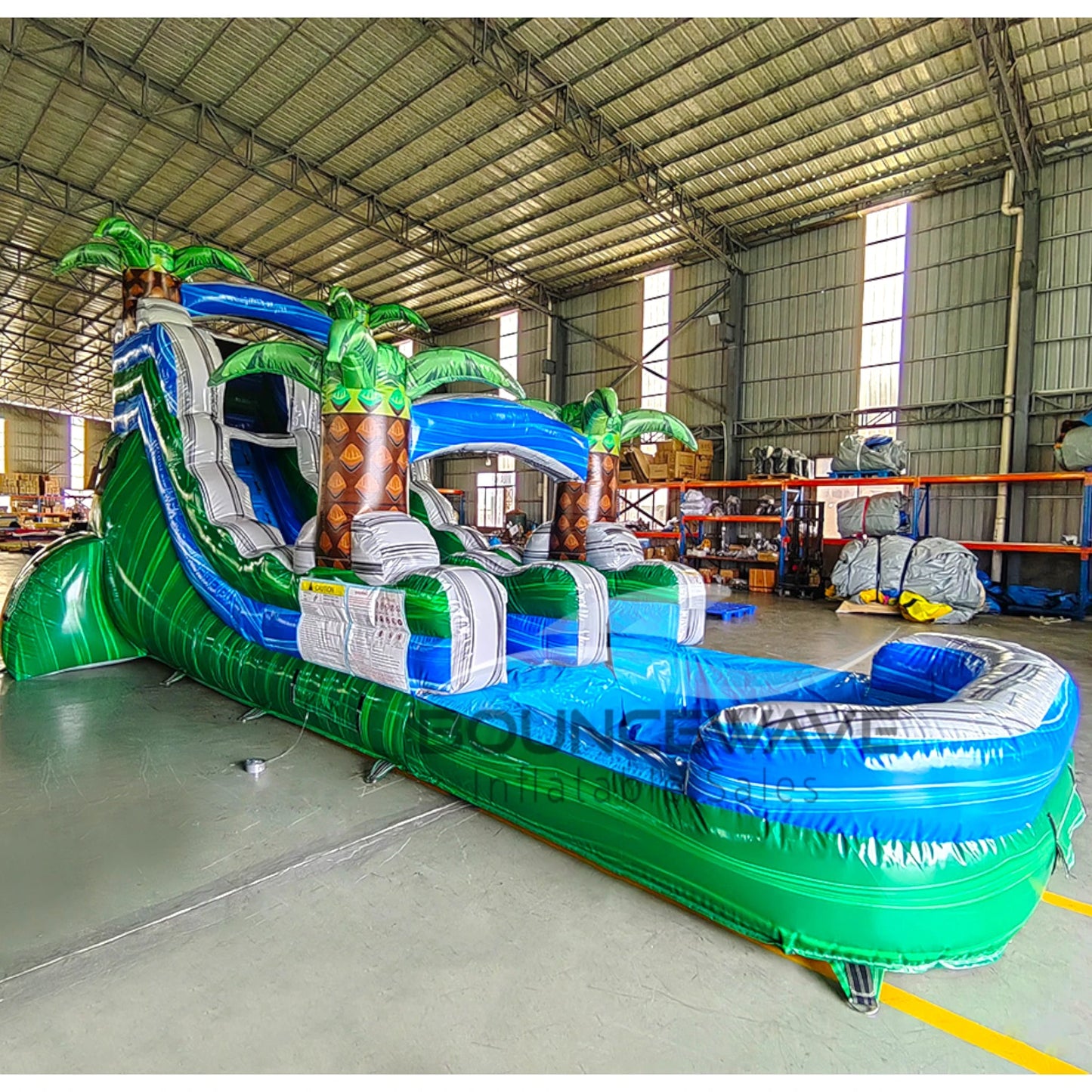 15ft Green Gush Commercial Single Lane Inflatable Water Slide side view displaying blue curved slide, green base, and inflatable palm tree decorations in indoor facility
