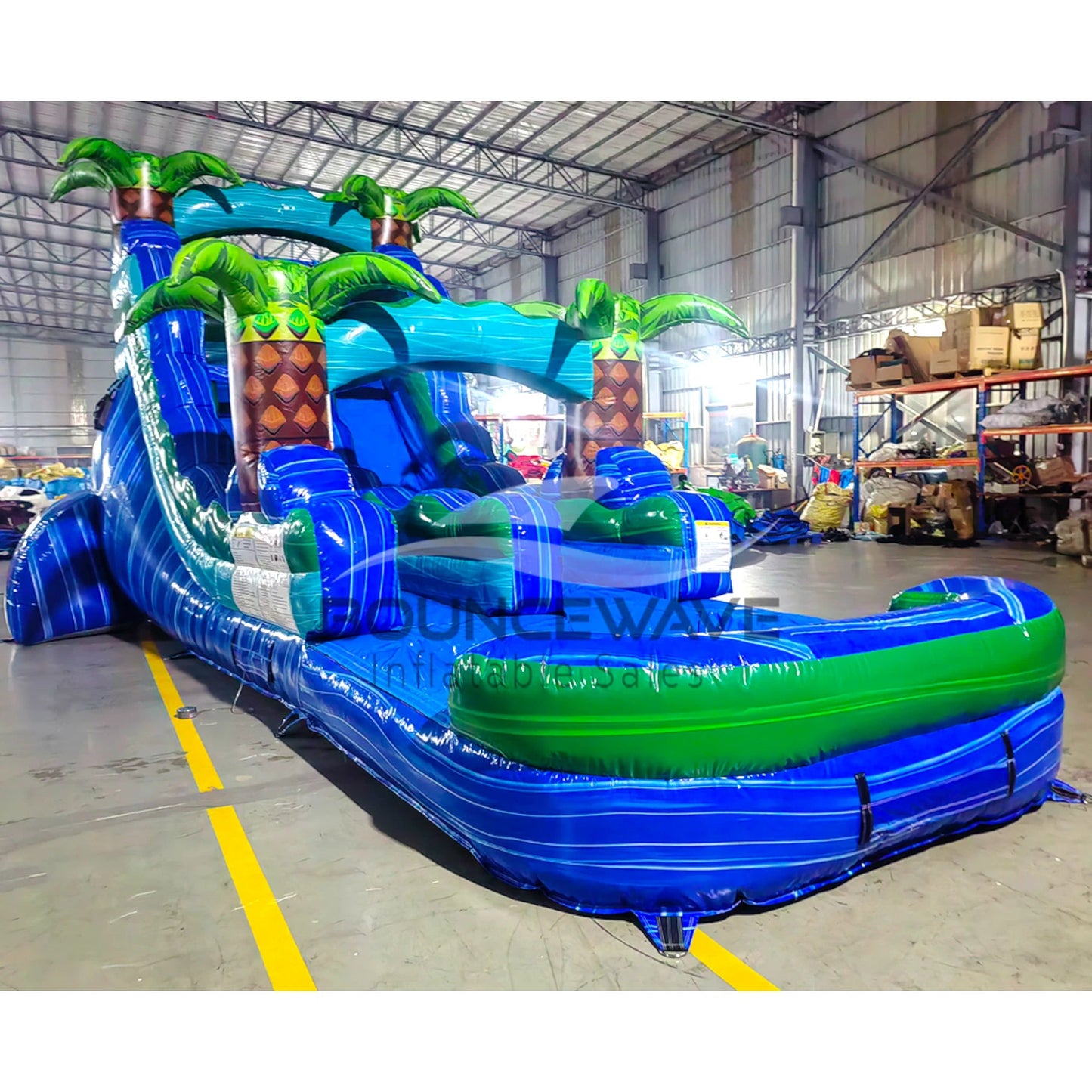 Large tropical-themed inflatable water slide featuring a single blue sliding lane, palm tree decorations, and an extended splash pool with wave-like design.