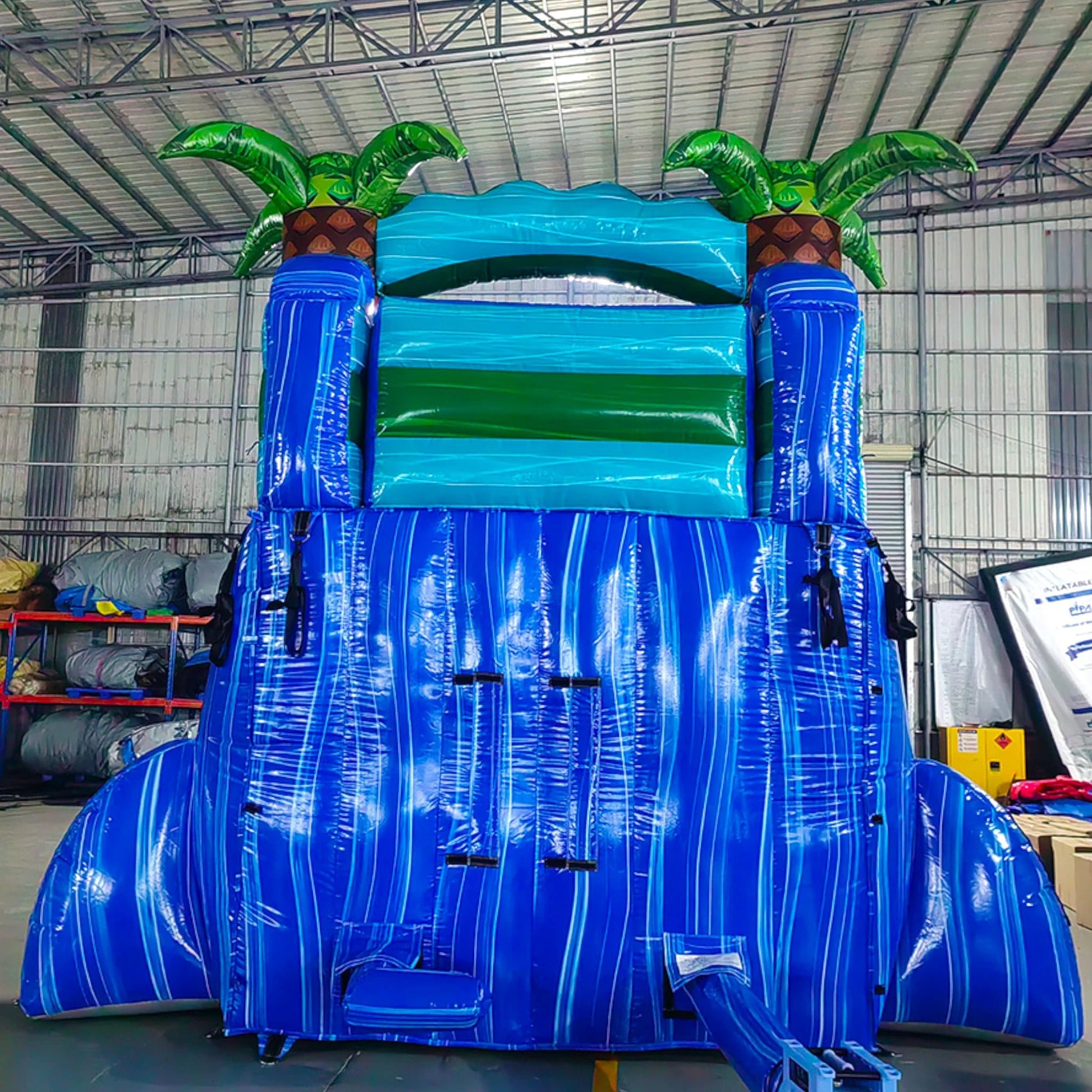 Rear view of Island Drop single lane water slide in indoor warehouse, showing tall blue structure with climbing area and palm tree decorations on top.