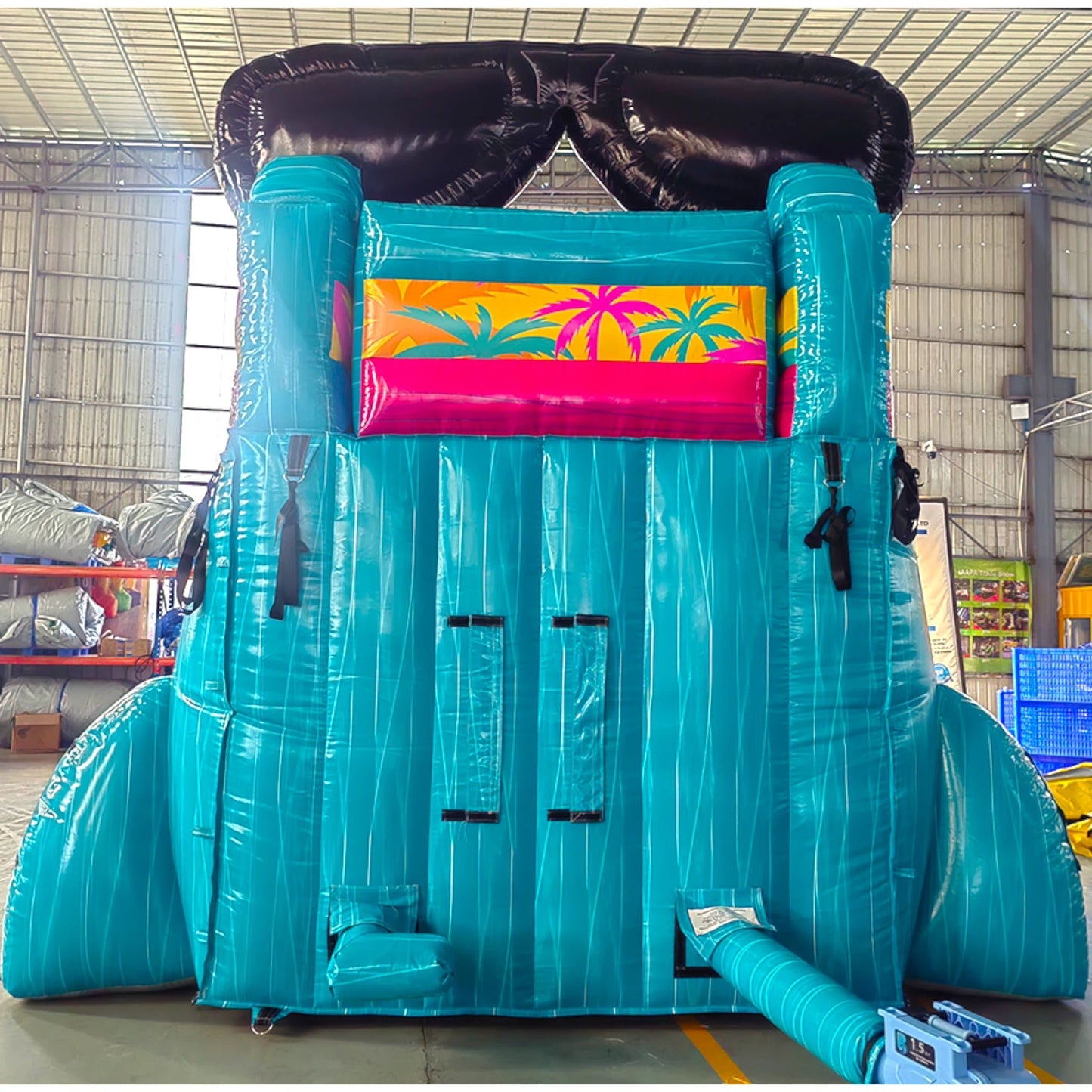 Rear view of tropical-themed inflatable water slide with large sunglasses design on top, showing tall turquoise structure and colorful palm tree decorations in indoor warehouse setting.