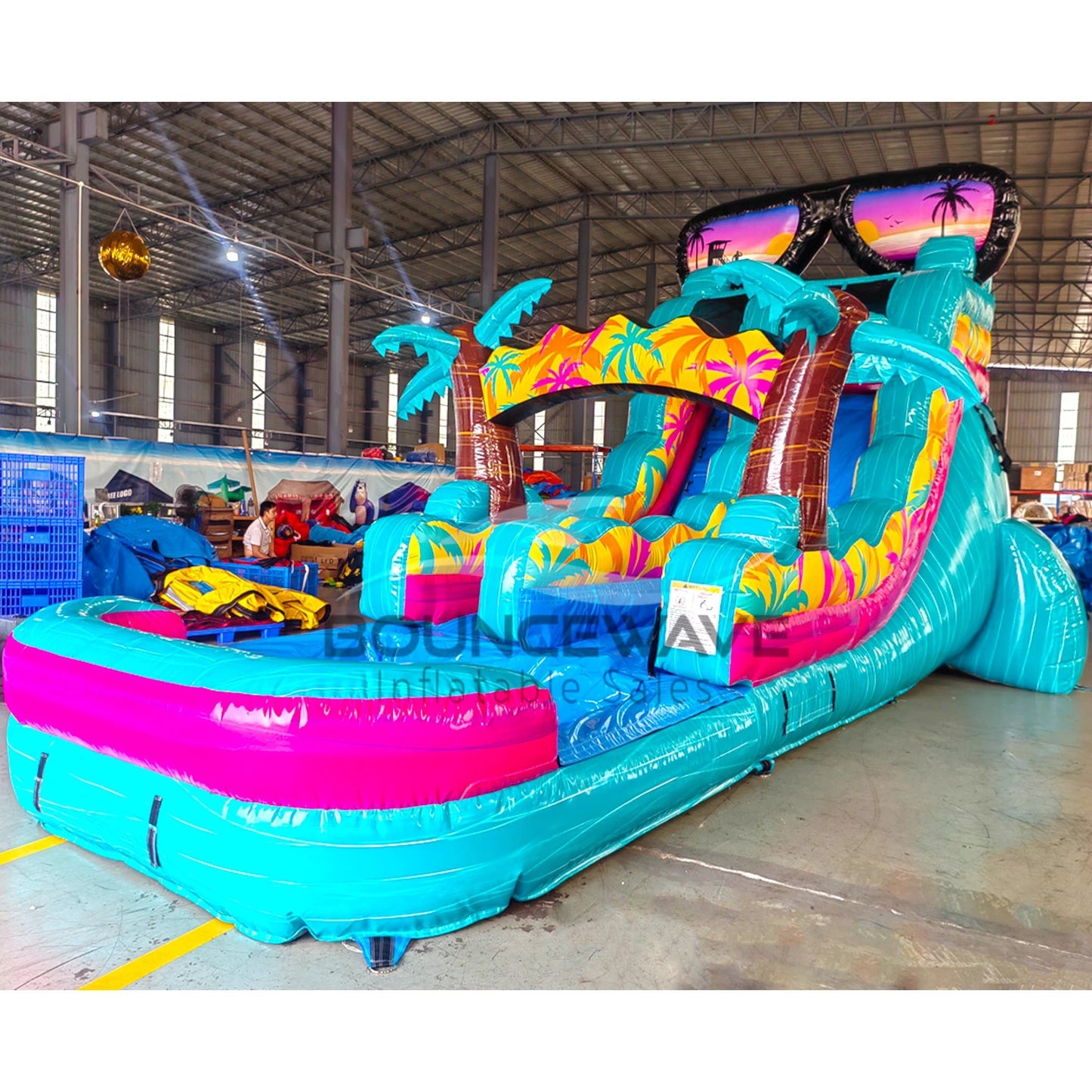 Front angled view of Island Flow inflatable water slide in indoor facility, showcasing sunglasses design, single blue sliding lane, palm trees, and large splash area with colorful tropical theme.