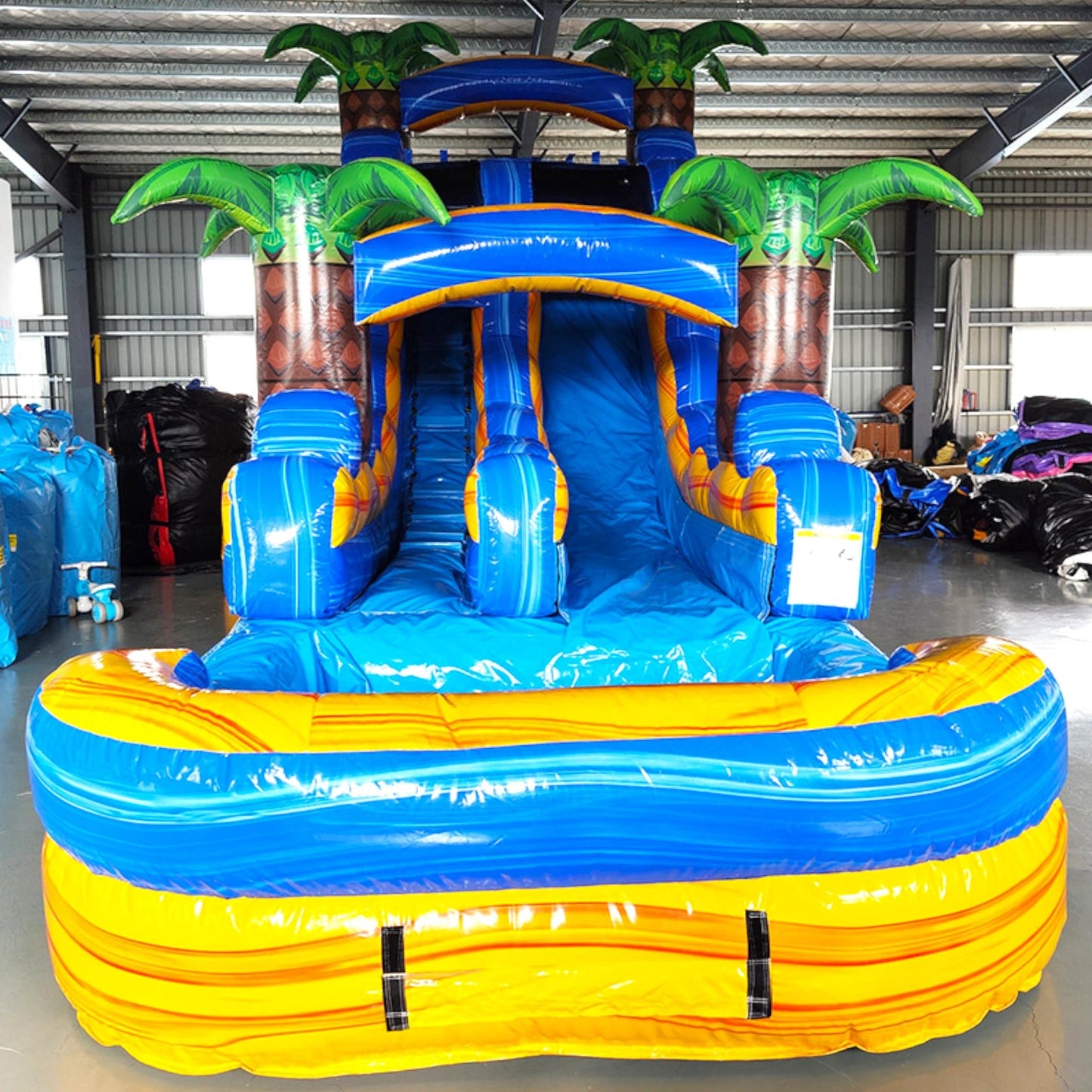 Front view of Lava Falls inflatable water slide displaying vibrant blue and yellow design, single lane, palm tree decorations, and spacious splash pool in indoor facility.