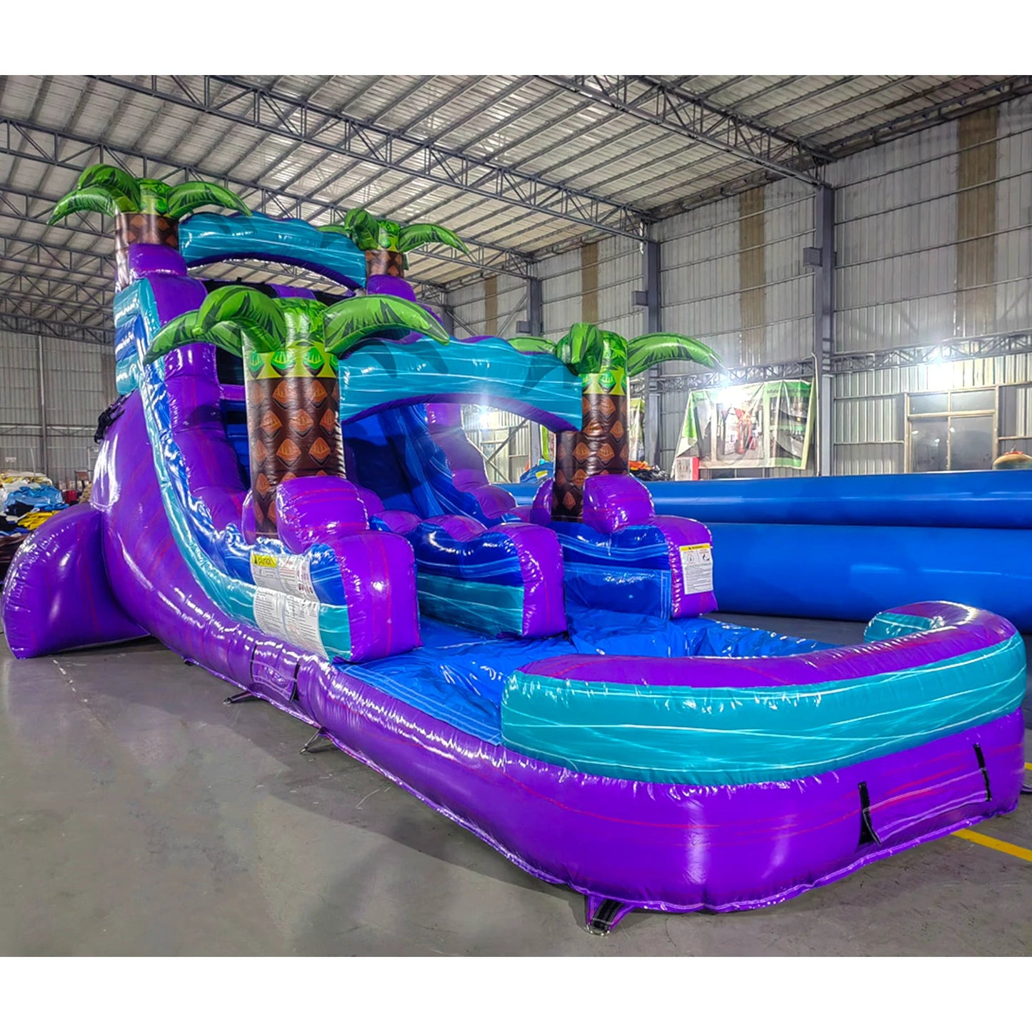 Large tropical-themed inflatable water slide featuring a single purple sliding lane, palm tree decorations, and an extended splash pool with vibrant blue and purple colors.