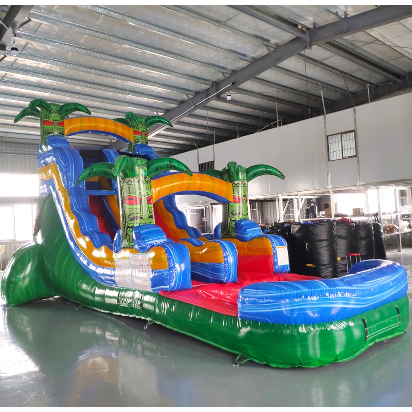 Large tropical-themed inflatable water slide featuring a single sliding lane, palm tree decorations, and an extended splash pool with vibrant blue, yellow, and green colors in indoor warehouse setting.