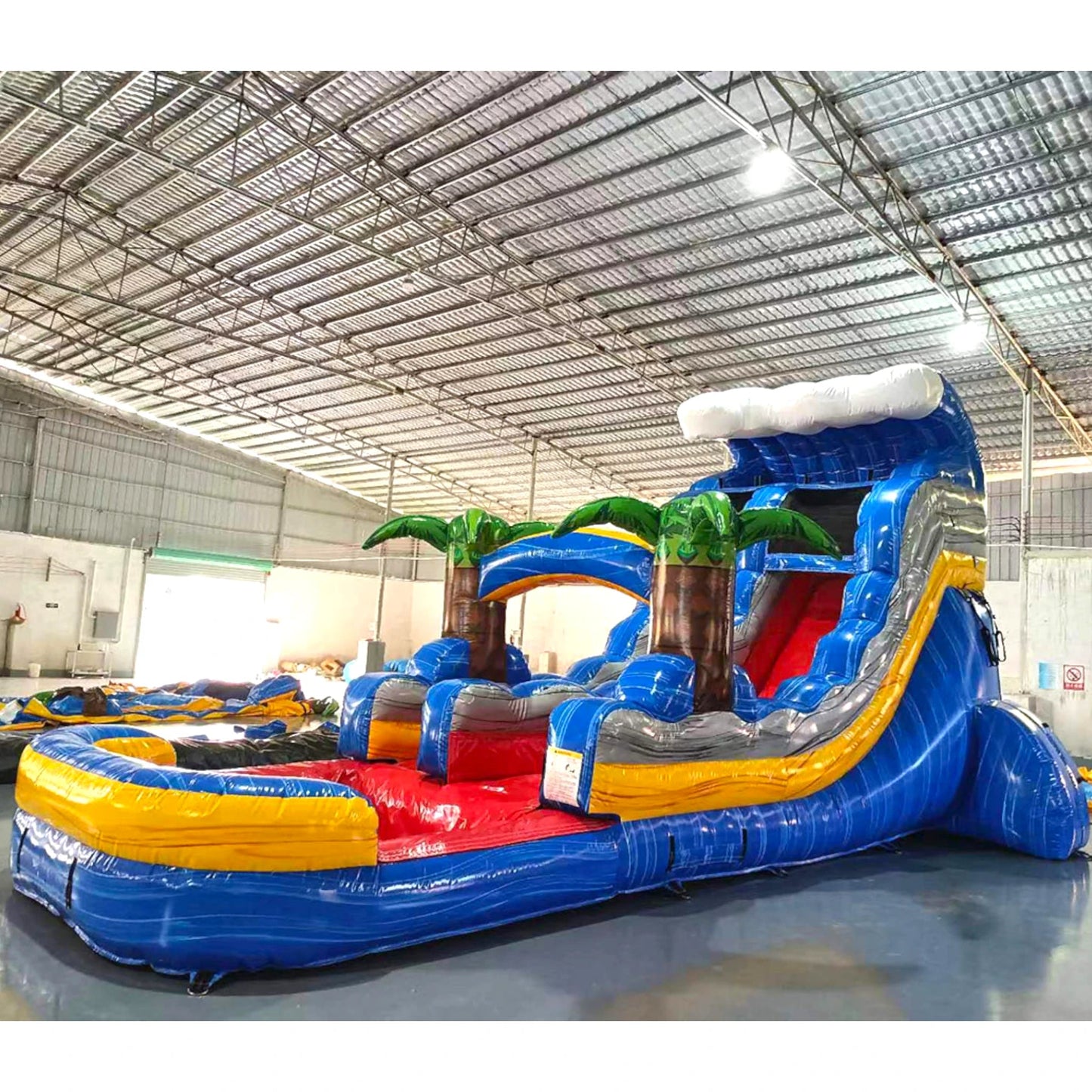 Large tropical-themed inflatable water slide featuring a single blue sliding lane, palm tree decorations, and an extended splash pool with wave-like design in outdoor setting.