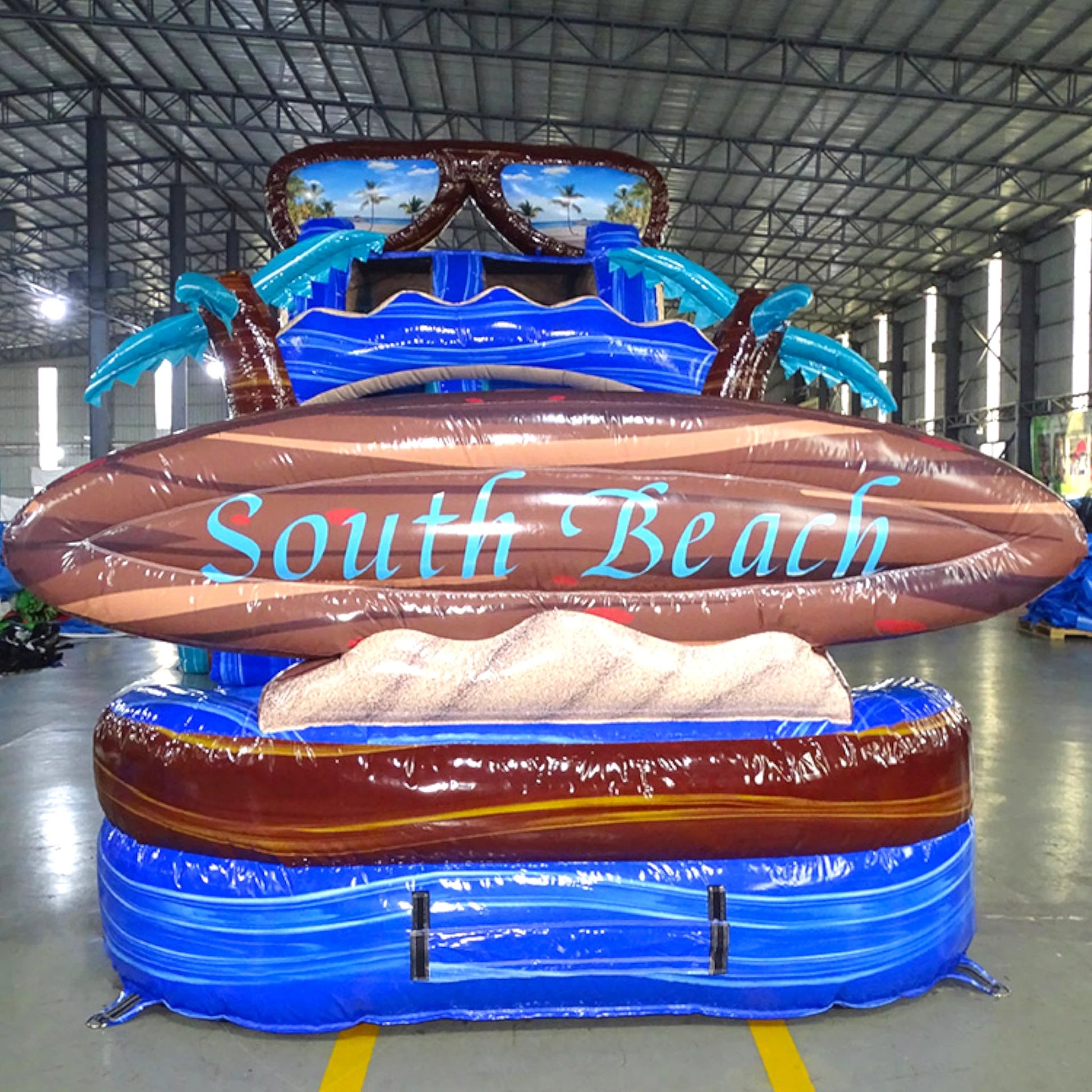 Front view of South Beach single lane water slide featuring sunglasses design on top, palm tree decorations, and "South Beach" sign on brown inflatable base.