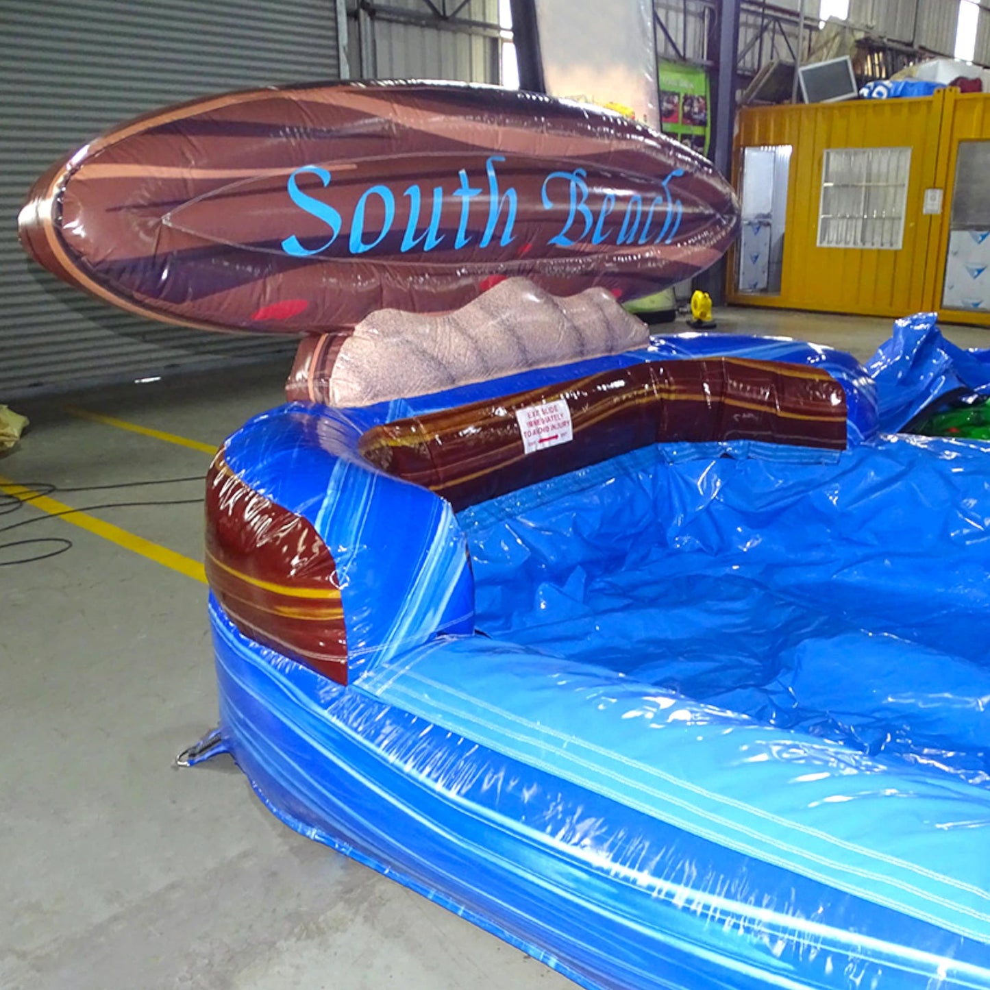 Inflatable blue and brown "South Beach" water slide with single lane and curved design, set up in an indoor warehouse space