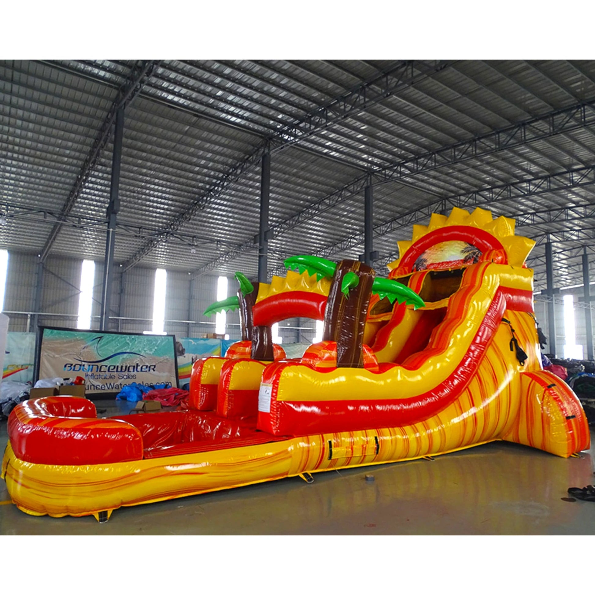 Side view of Summer Sizzler single lane water slide in indoor warehouse, showing full length with yellow sliding surface, sun-shaped top, palm trees, and large splash area.