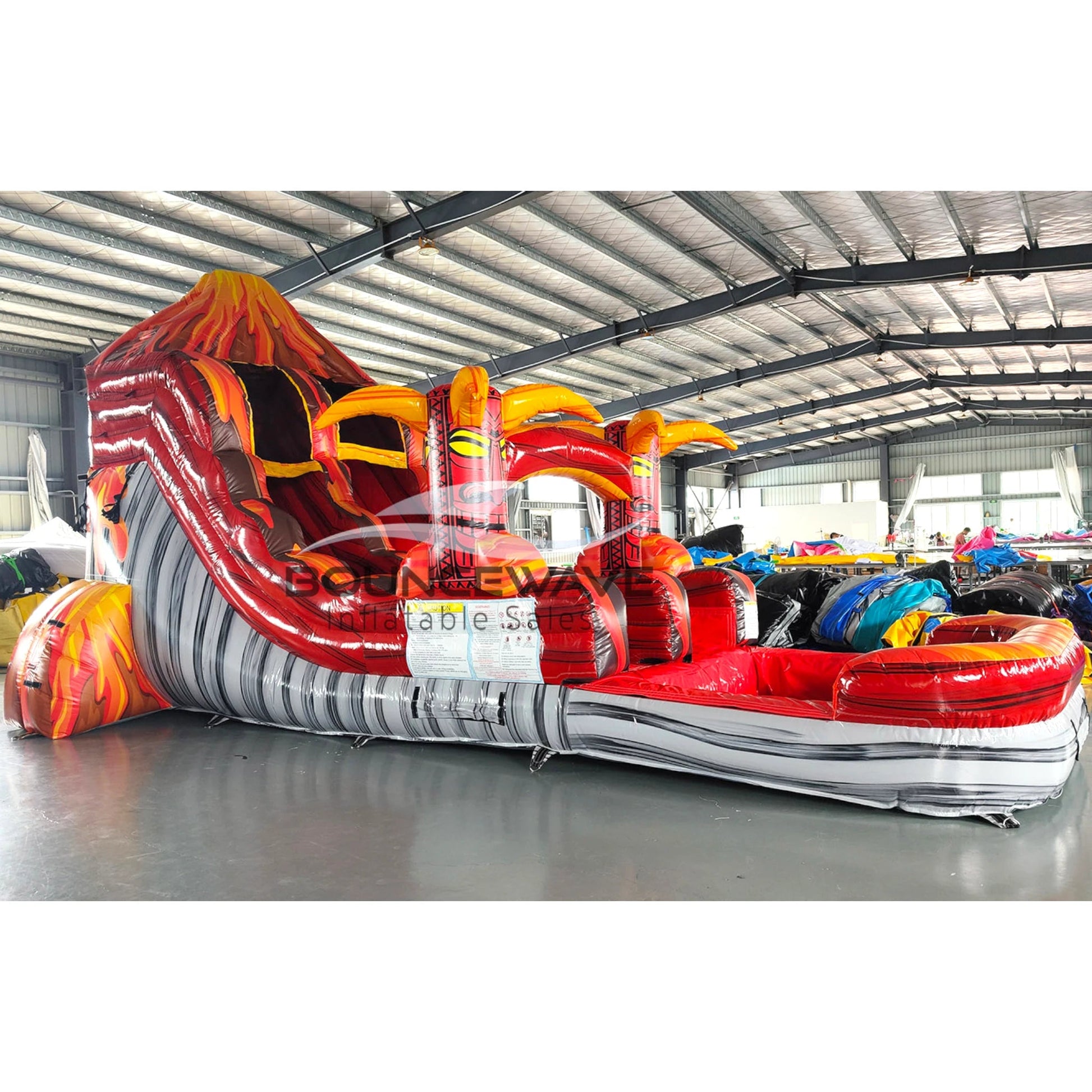 Large volcano-themed inflatable water slide featuring a single red sliding lane, tiki mask decorations, and an extended splash pool with lava-like design in indoor warehouse setting.