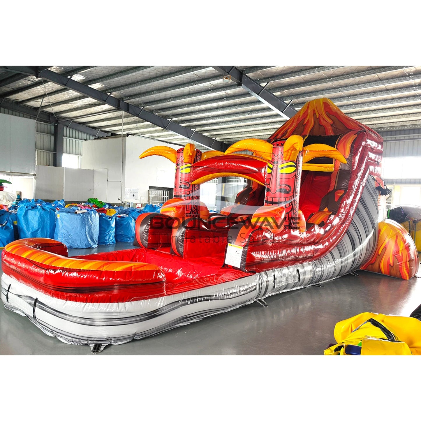 Side view of Tiki Volcano single lane water slide showing full length with red sliding surface, tiki mask accents, and large splash area with flame-inspired colors.