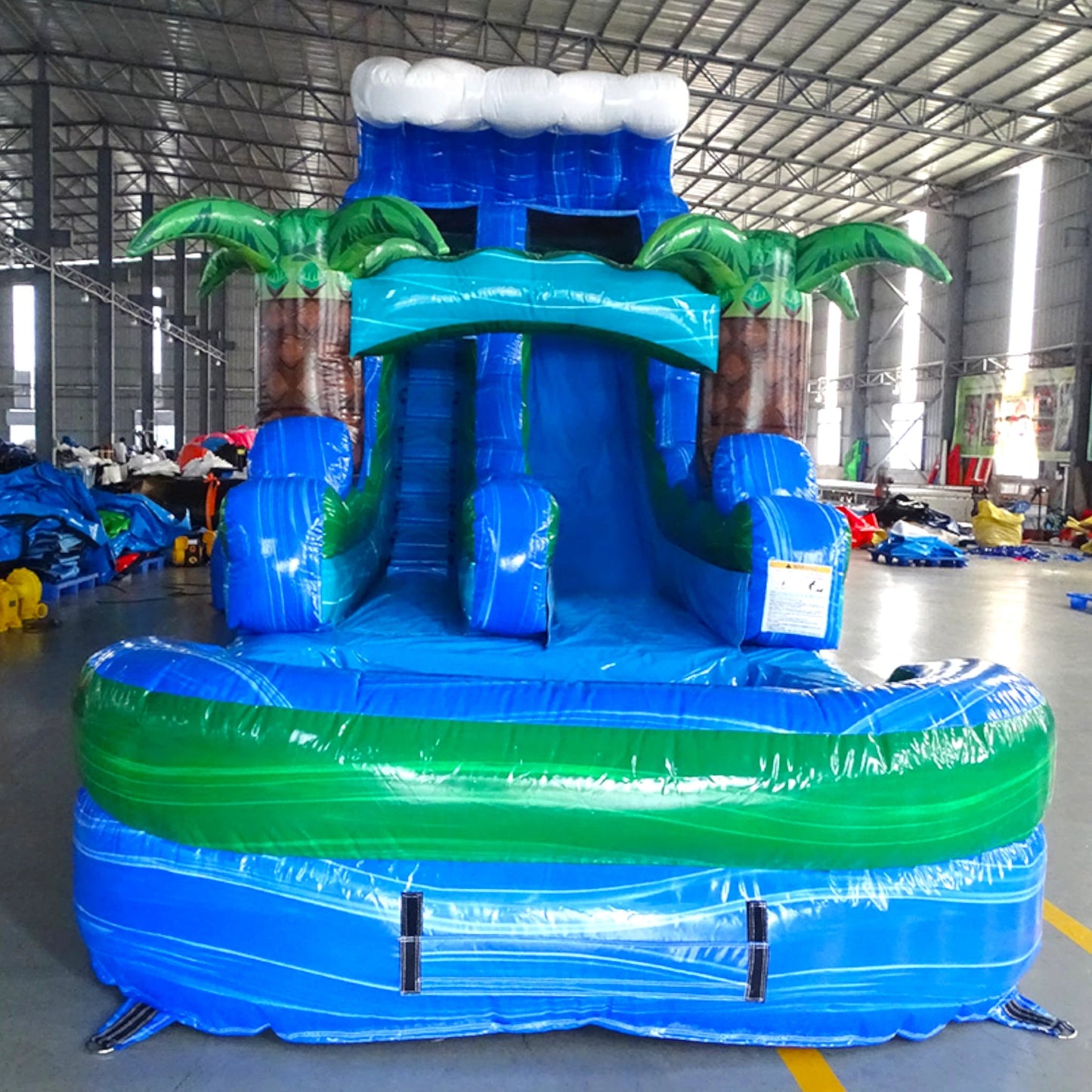 Front view of Trop Wave inflatable water slide displaying vibrant blue design, single lane, palm tree decorations, and spacious splash pool with green rim in indoor facility.