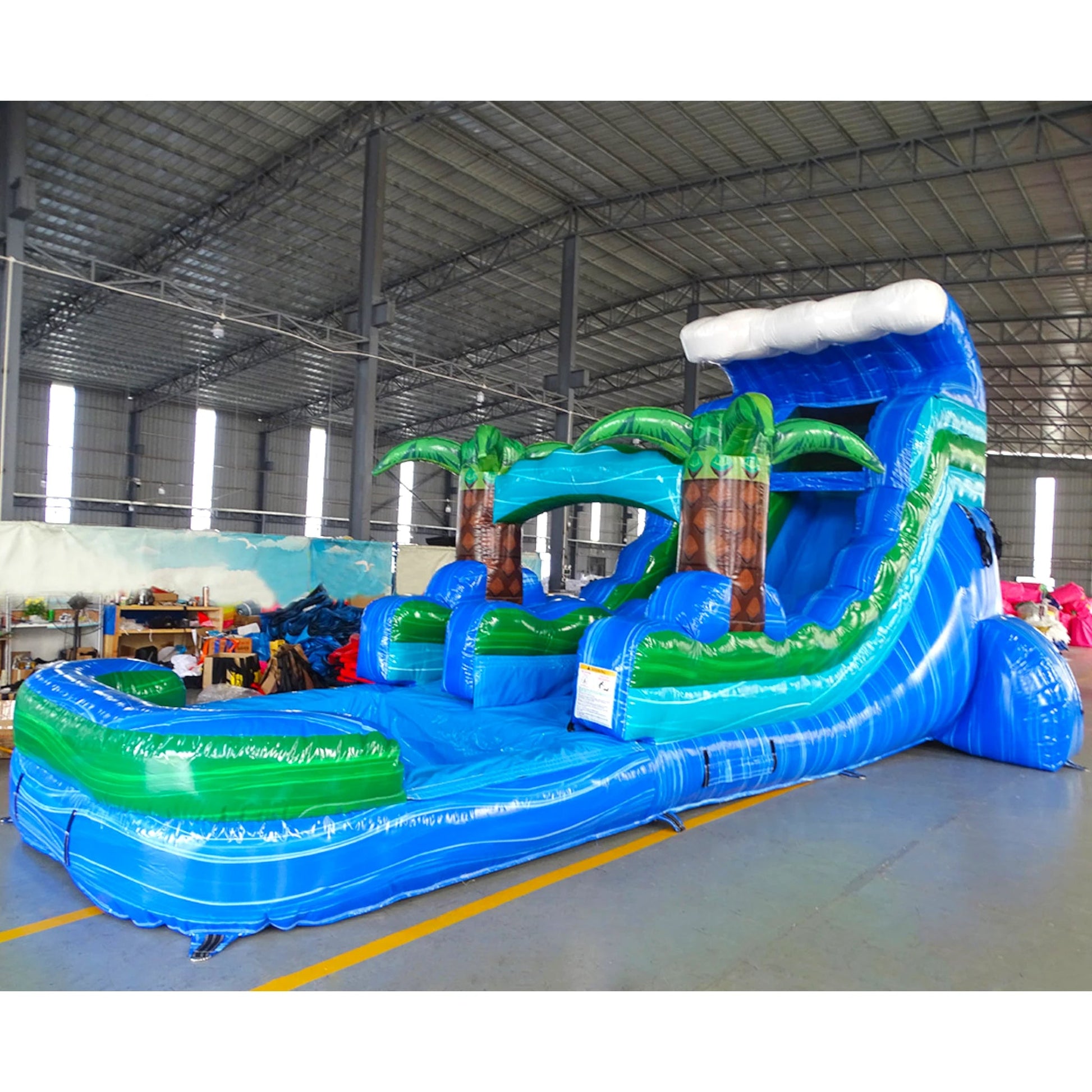 Large tropical-themed inflatable water slide featuring a single blue sliding lane, palm tree decorations, and an extended splash pool with wave-like design in indoor warehouse setting.