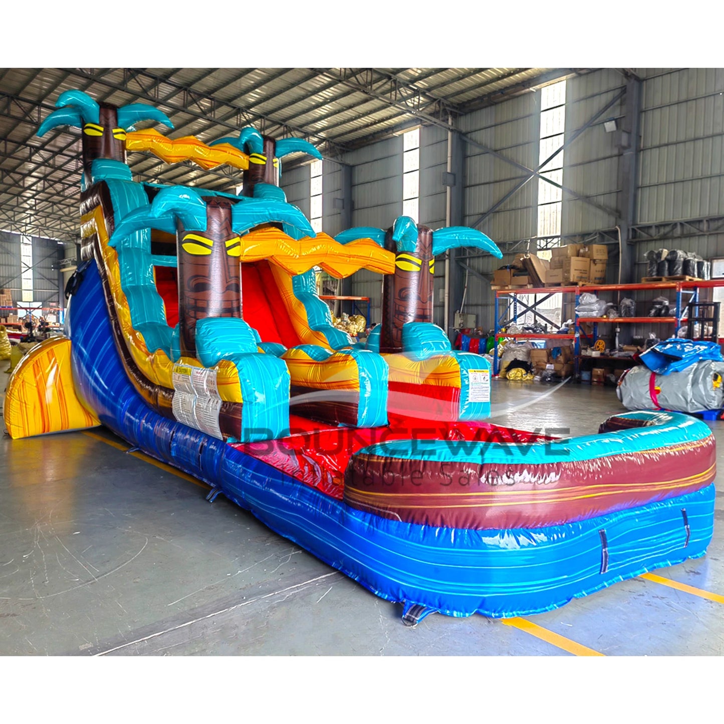 Large tropical-themed inflatable water slide featuring a single blue sliding lane, palm tree decorations, and an extended splash pool with vibrant blue and yellow colors in indoor warehouse setting.