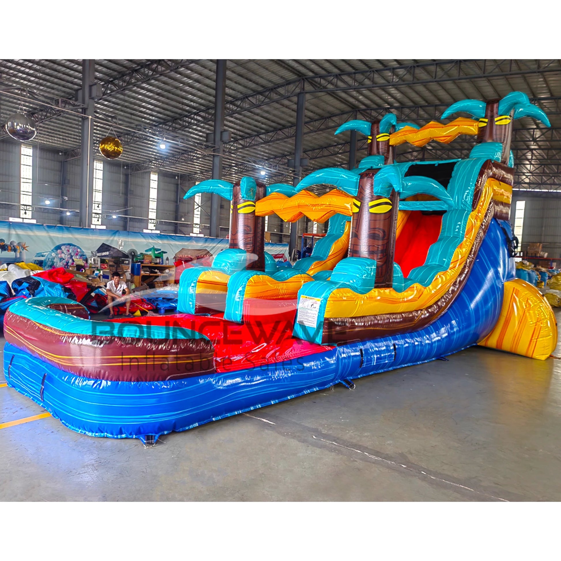 Large tropical-themed inflatable water slide featuring a single blue sliding lane, palm tree decorations, and an extended splash pool with vibrant blue and yellow colors in indoor warehouse setting.