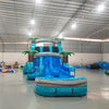 Large inflatable water slide with tropical theme, featuring blue sliding lanes, palm tree decorations, and a colorful splash pool area. The slide combines open and enclosed sections, set up in an indoor warehouse with other inflatables visible.