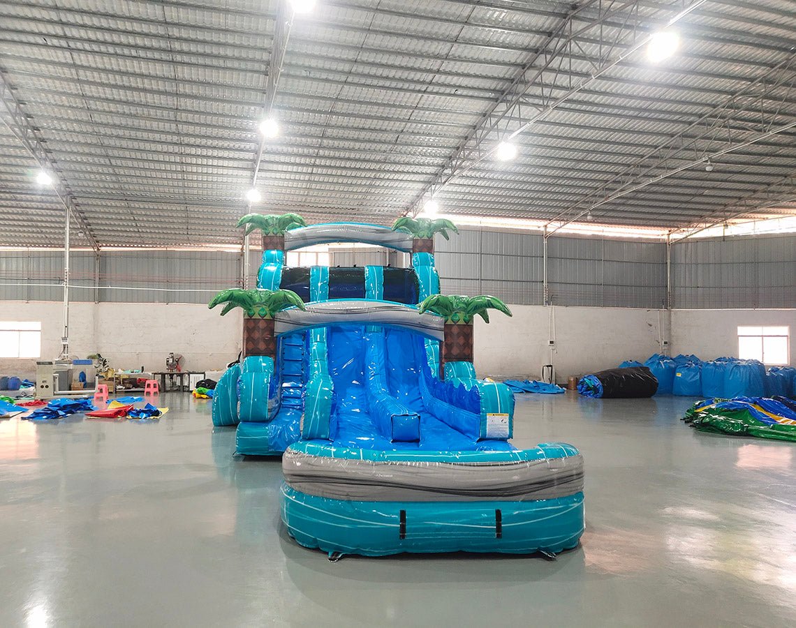 Large inflatable water slide with tropical theme, featuring blue sliding lanes, palm tree decorations, and a colorful splash pool area. The slide combines open and enclosed sections, set up in an indoor warehouse with other inflatables visible.