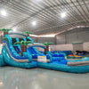 Front view of the 16' Bahama Blast Hybrid DL inflatable water slide, showcasing its blue sliding lanes, palm tree decorations, and spacious splash pool. The slide combines open and tube slide elements, set up in an indoor warehouse with other equipment visible.