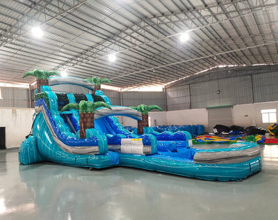 Front view of the 16' Bahama Blast Hybrid DL inflatable water slide, showcasing its blue sliding lanes, palm tree decorations, and spacious splash pool. The slide combines open and tube slide elements, set up in an indoor warehouse with other equipment visible.