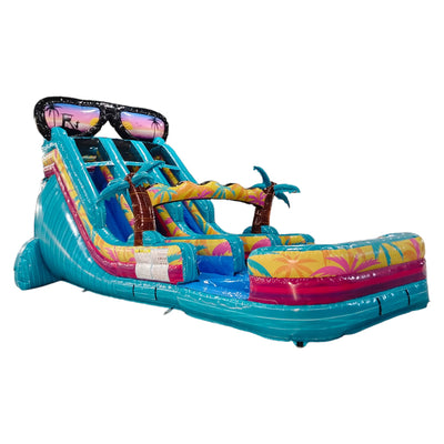 Large inflatable water slide with tropical theme, featuring a giant sunglasses decoration on top, three blue sliding lanes, palm tree inflatables, and a colorful splash pool area, set up in an indoor warehouse.