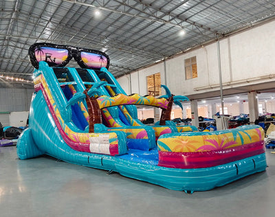 Front angled view of the 18ft Island Flow Center Climb water slide, showcasing its three blue sliding lanes, tropical-themed decorations including palm trees and giant sunglasses, and a colorful splash pool, set up in an indoor warehouse with other inflatables visible in the background.