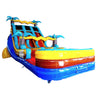 Large inflatable water slide with tropical theme, featuring blue sliding lanes, palm tree decorations, and a colorful splash pool area. The slide has a white foam-like top and is set up in an indoor warehouse with other inflatables visible in the background.