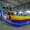 Side view of 18ft Tropic Shock Center Climb water slide showing vibrant blue sliding lanes, inflatable palm trees, and a large green and blue splash pool area. The slide is set up in an indoor facility with metal roofing and other equipment visible.