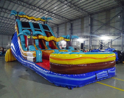 Side view of 18ft Tropic Shock Center Climb water slide showing vibrant blue sliding lanes, inflatable palm trees, and a large green and blue splash pool area. The slide is set up in an indoor facility with metal roofing and other equipment visible.