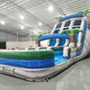 Three-quarter view of 18ft Aloha Splash water slide displaying blue lanes, green accents, inflatable palm trees, central climbing structure, and spacious splash area with tropical-themed design elements.