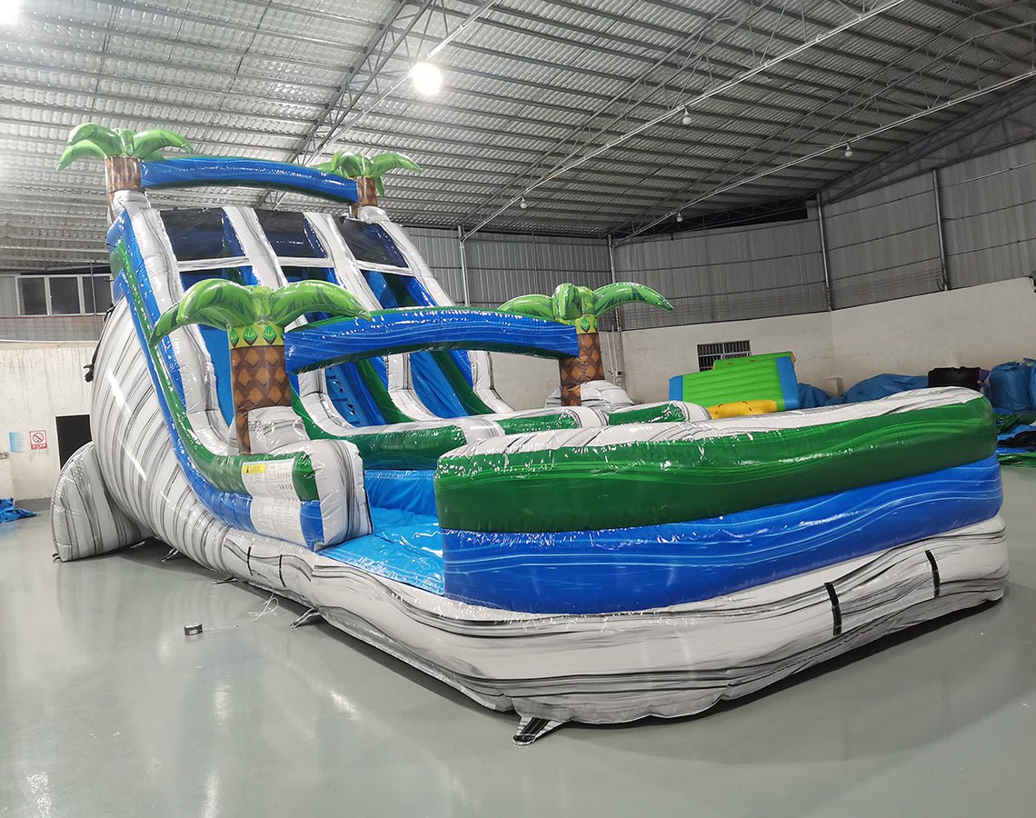 Three-quarter view of 18ft Aloha Splash water slide displaying blue lanes, green accents, inflatable palm trees, central climbing structure, and spacious splash area with tropical-themed design elements.