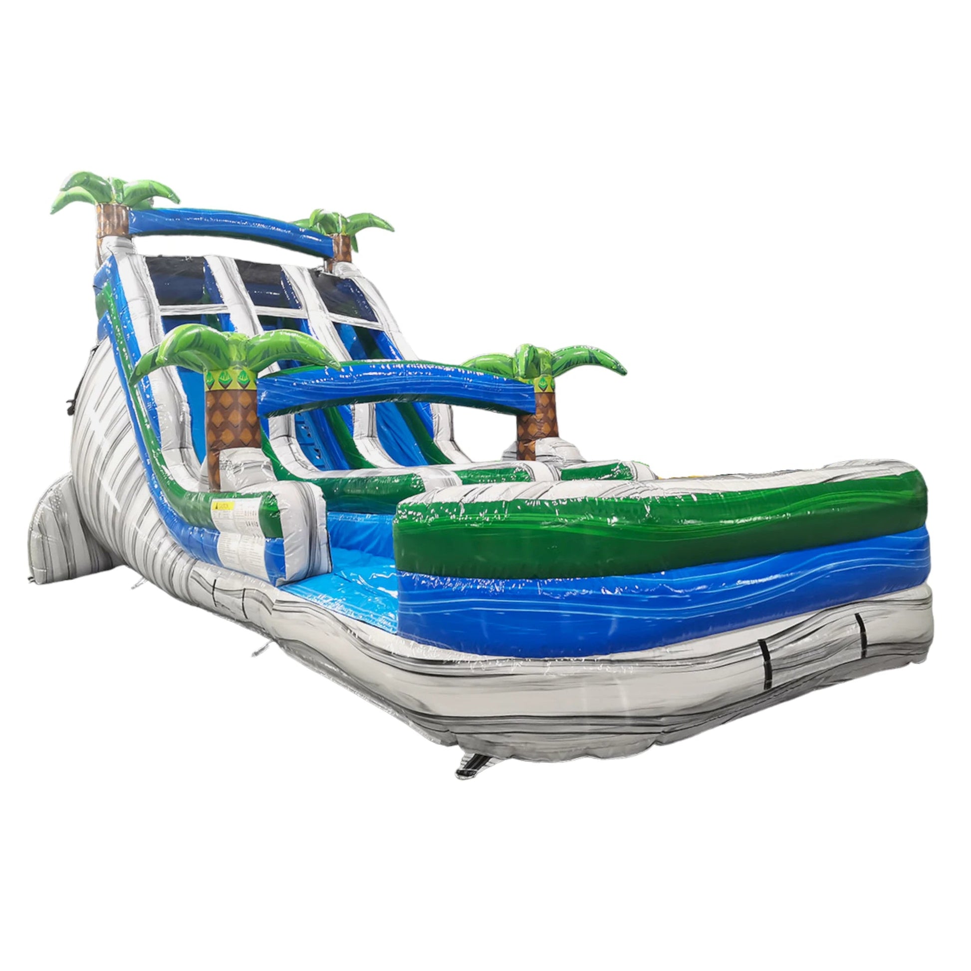 Side view of 18ft Aloha Splash inflatable water slide featuring three blue lanes, tropical palm tree decorations, central climbing structure, and large splash pool with green and blue accents, set up in an indoor warehouse.