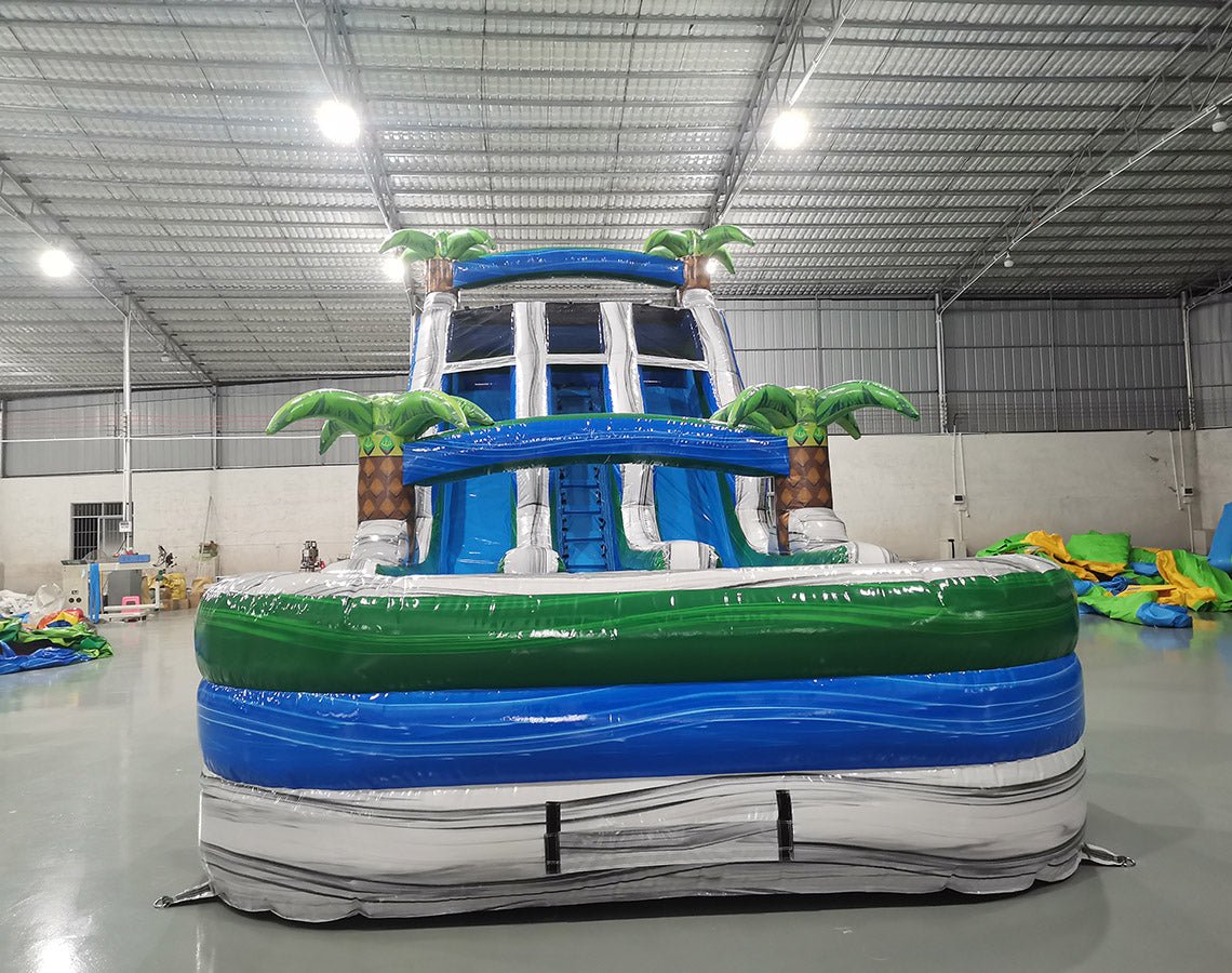 Front view of 18ft Aloha Splash inflatable water slide featuring three blue lanes, tropical palm tree decorations, central climbing structure, and large splash pool with green and blue accents.